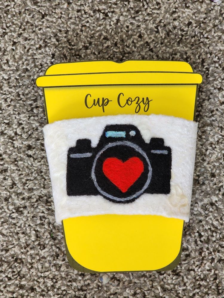 White wet felted cup cozy with a needlefelted camera and heart in the center of the camera lens on the front of the cozy displayed on a cardboard cup.