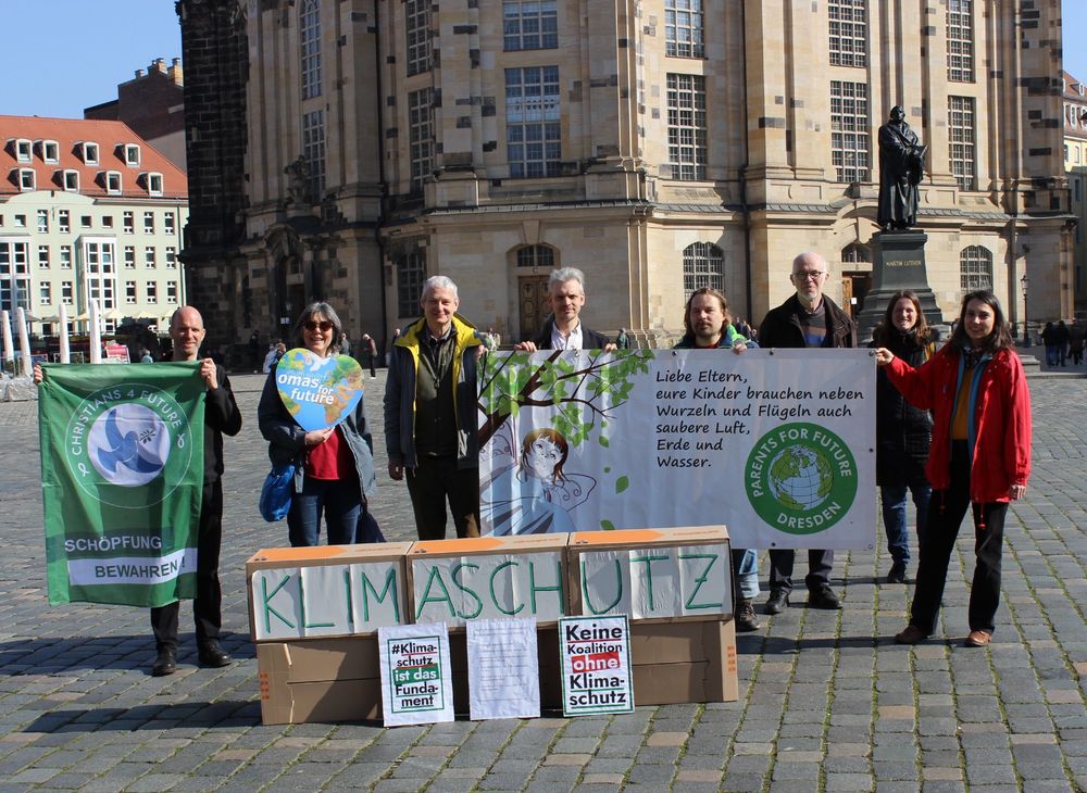 Menschen stehen mit Bannern und Fahnen vor der Frauenkirche. Sie sind von den Organisationen Parents For Future, Christians For Future, Omas For Future und Extinction Rebellion. Sie fordern Klimaschutz und die Bewahrung der Schöpfung.