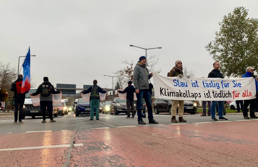 Menschen blockieren eine Straße an der TU Dresden. Sie halten ein Banner „Stau ist lästig für Sie; Klimakollaps ist tödlich für alle“