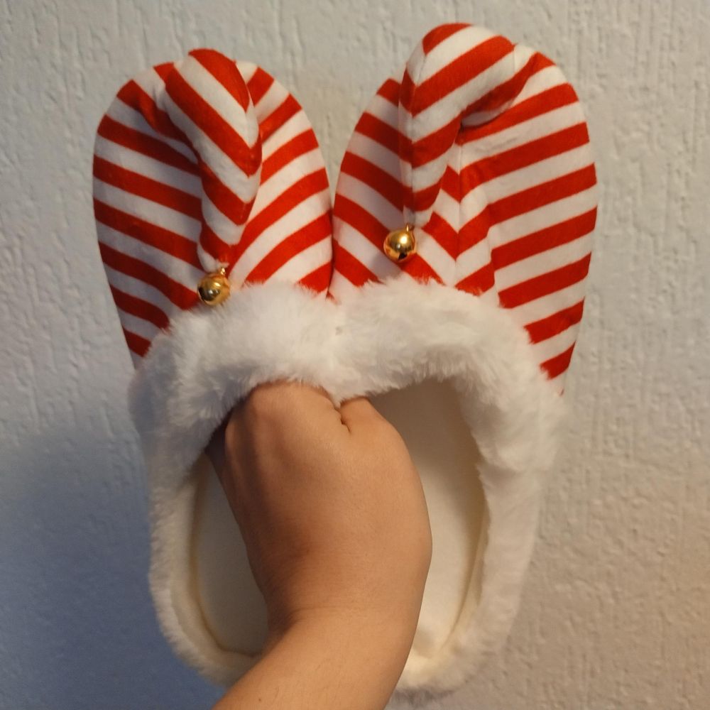 Eine Hand hält ein Paar auffällige, weihnachtlich designte Hausschuhe vor eine helle Wand. Die Puschen sind rot-weiß gestreift wie Zuckerstangen, haben flauschige, weiße Kunstfellränder und an der Spitze jeweils ein kleines goldenes Glöckchen. Die Form erinnert an Elfen- oder Weihnachtsmannschuhe, verspielt und cozy zugleich.