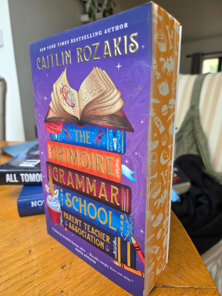 Paperback for Caitlin Rozakis' The Grimoire Grammar School Parent Teacher Association. Cover depicts a tower of tomes, with the one on top open on pages decorated with a circle pentagon and other scribblings. On the bottom, a unicorn cupcake. The paperback has orange painted edges, with Halloween-ish symbols (skulls, cauldron, cupcake, unicorn, bone, candle, etc). It's super cute and super orange and purple. I love it.