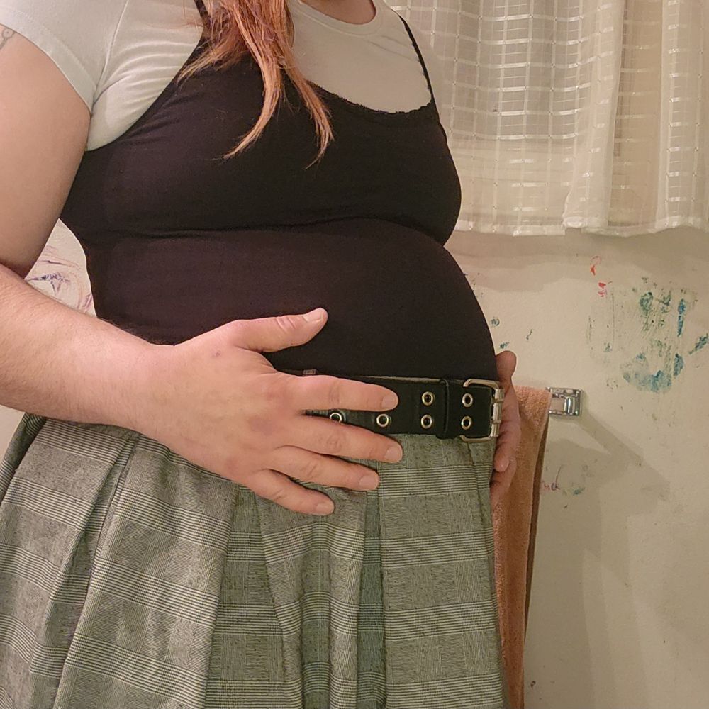 Me, wearing a black camisole over a white t-shirt with a grey plaid skirt and a black grommet belt. My belly is noticeably bloated and resembles a pregnant belly.
