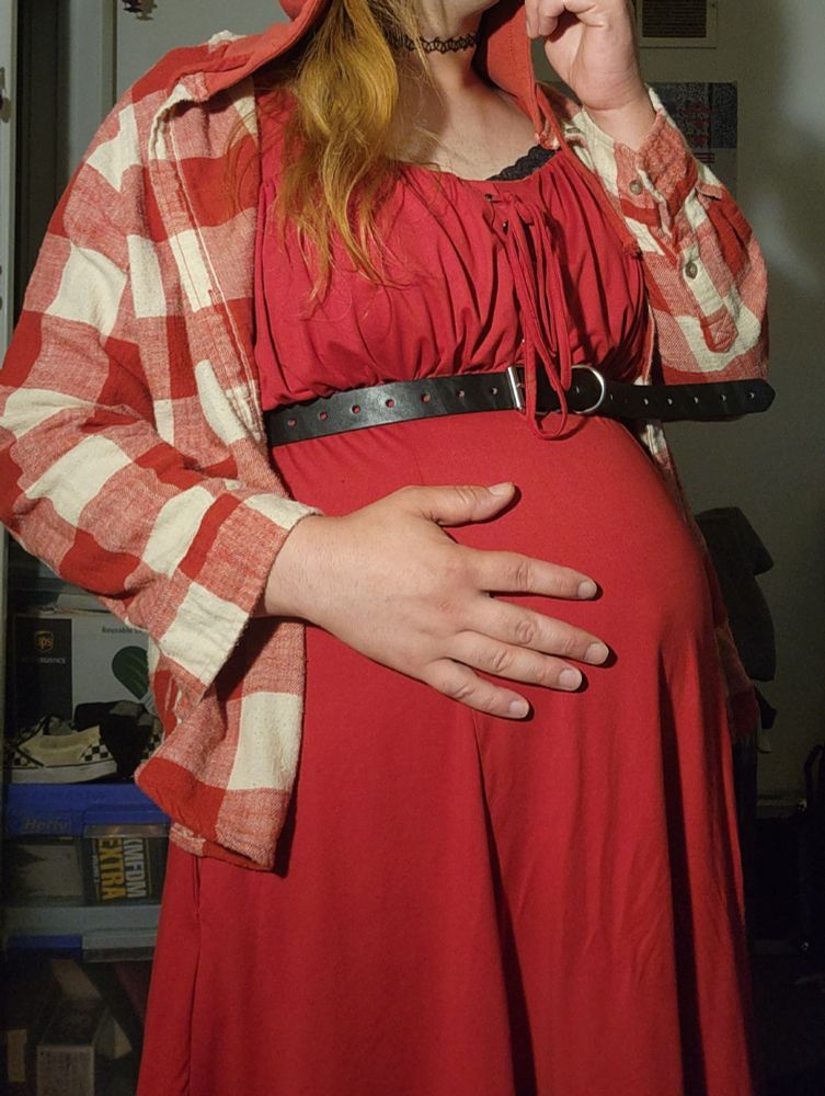 Me dressed as Little Red Riding Hood with a bloated belly after eating the wolf, wearing a red dress with black belt and plaid coat.