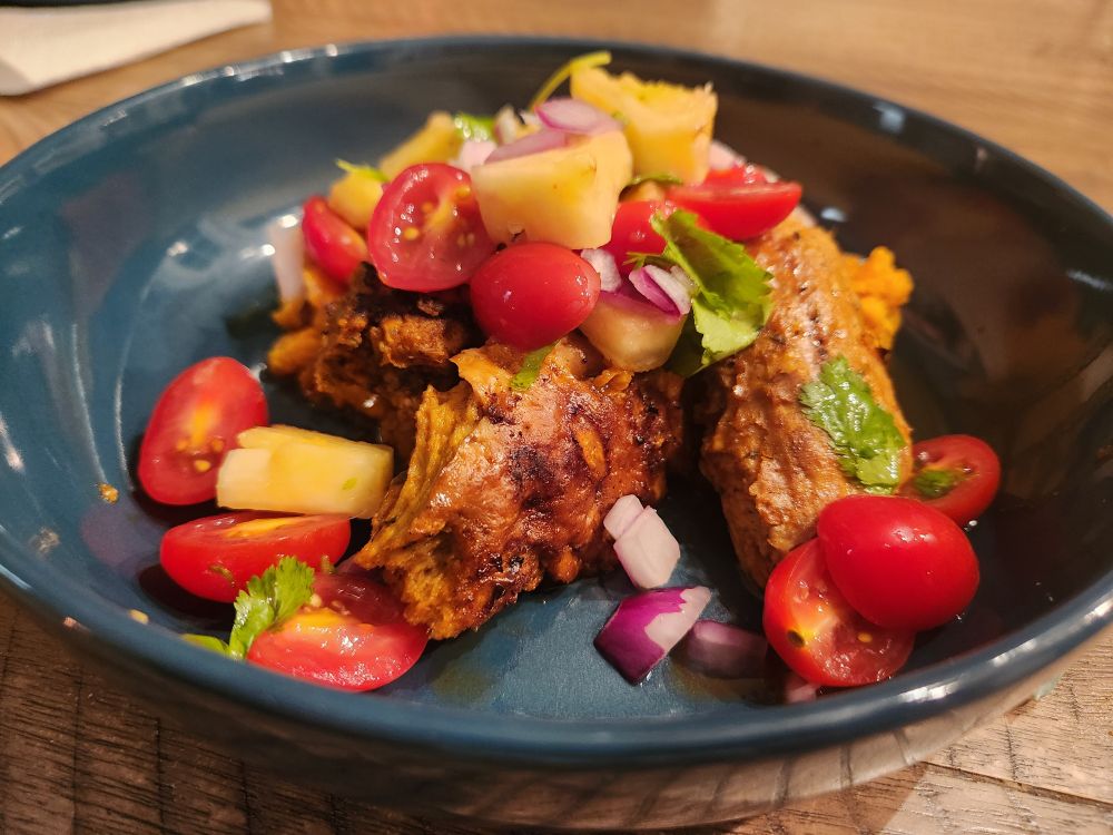 A colorful dark teal plate showing grilled seasoned vegan chik'n, mashed sweet potatoes, and a pineapple salsa with cherry tomatoes, pineapple, red onion, cilantro, and lime. 