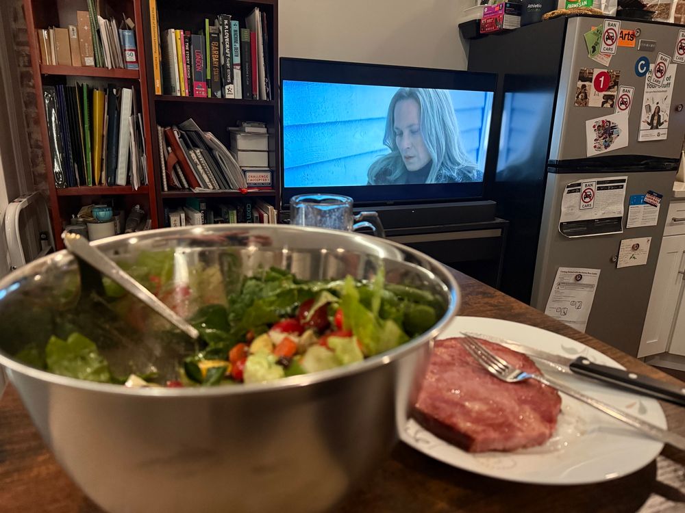 A large salad and a hamsteak in the foreground. Bookshelves, fridge, and a television of Patricia Arquette playing Harmony Cobel in the show Severance. 