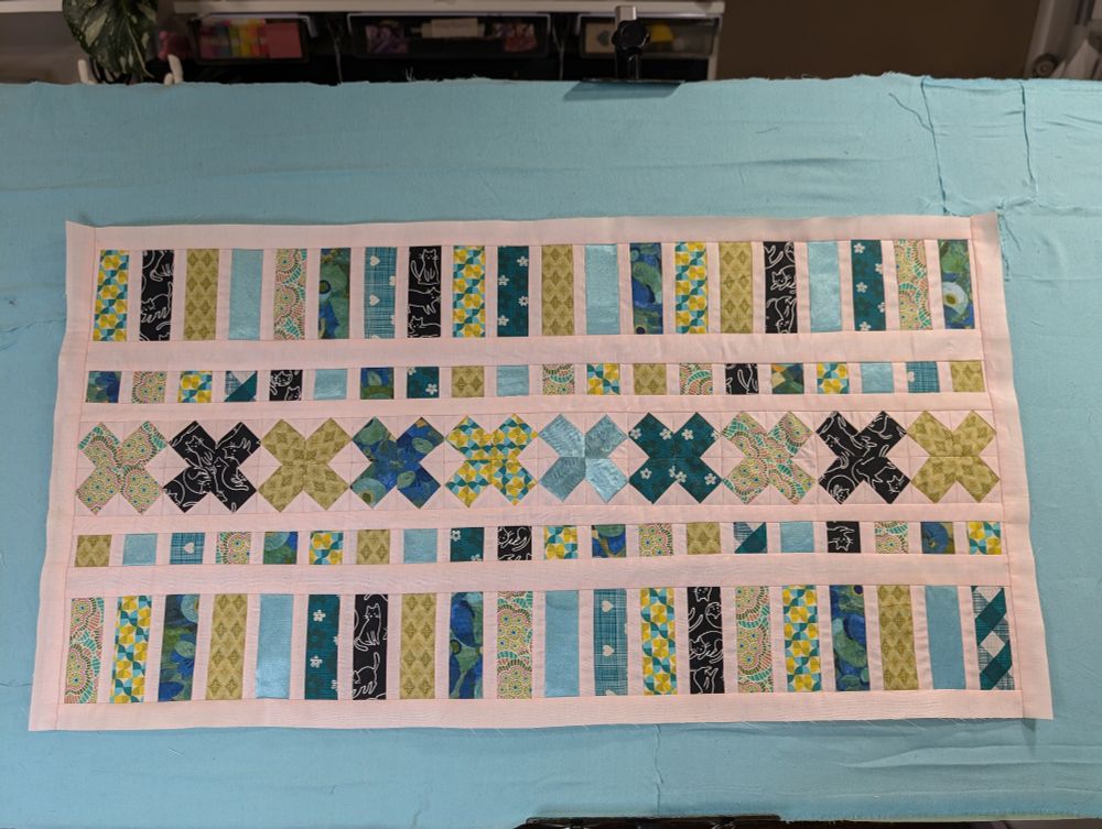 Photo of a finished quilt top of X's, strips and squares in shades of teal, aqua, and greens with a pale pink background.