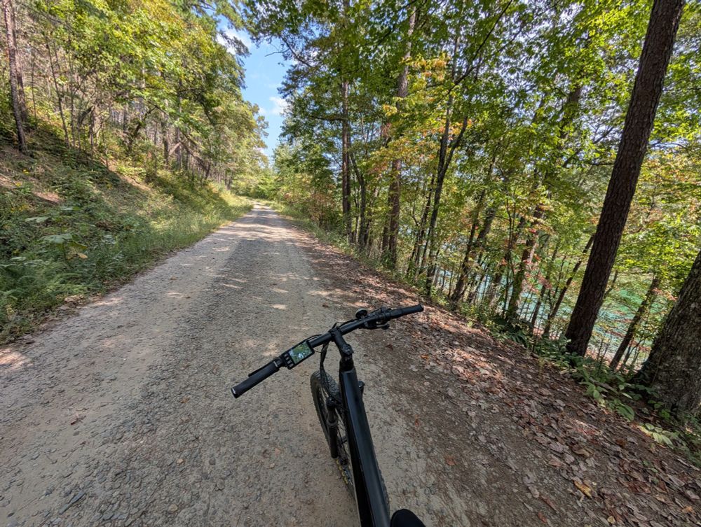 Gravel forest service road, trees on both sides with early fall colors, a lake can be seen thru the trees on the right.  Bike in foreground.