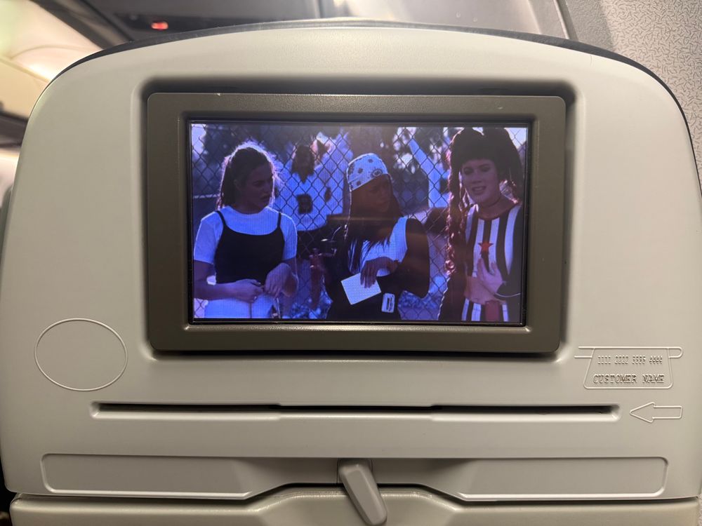 A seat back on a flight, showing a tv (displaying a frame from the show clueless with three high school students) and a credit card reader 