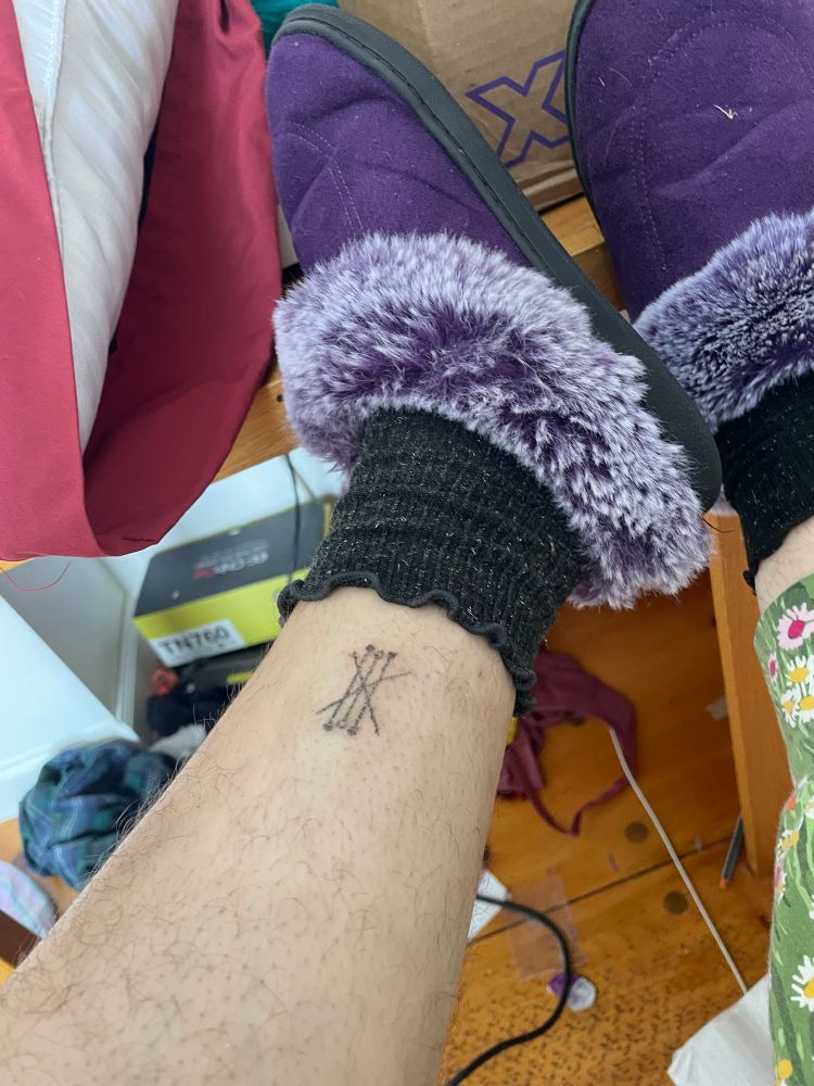 a pale, hairy leg wearing a black sock and a purple fuzzy slipper. on the ankle is a raw-looking stick-and-poke tattoo composed of three parallel black lines crossed by two more in an x. the top and bottom parallel lines end in filled-in circles while the middle line ends in open circles. 