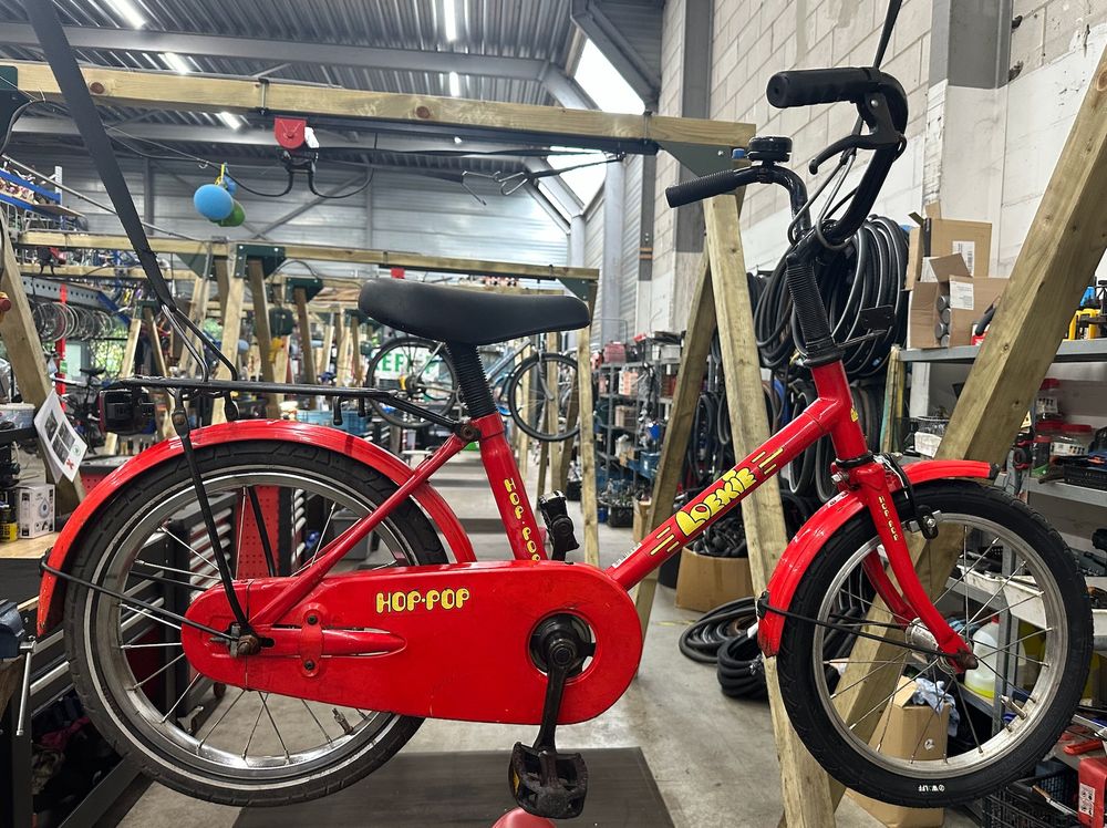 Bright red child's bike hanging from a bike lift in the BikeFlip Utrecht workshop. The brand is Loekie and the type is Hop-Pop. / Felrode kinderfiets hangt aan een fietslift in de werkplaats van BikeFlip Utrecht. Het merk is Loekie en het type is Hop-Pop.