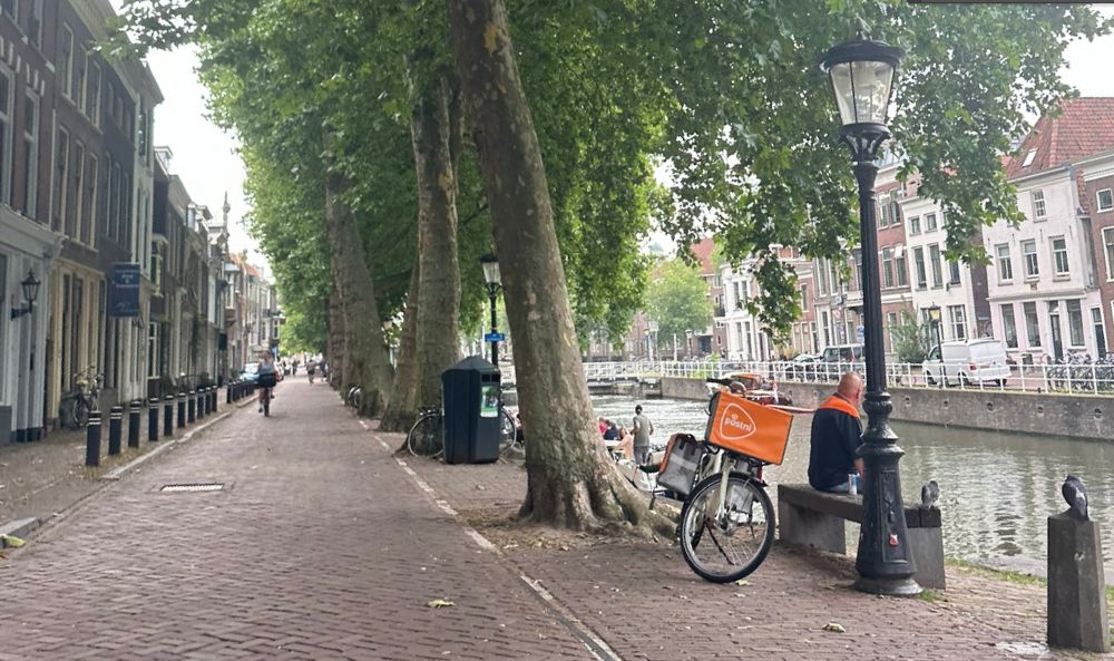 A post deliverer sits on a bench along a canal under large trees. Cobbled roads are lined  with 2 and 3-story buildings.
Een postbezorger zit op een bankje langs een kanaal onder grote bomen. Langs geplaveide wegen staan gebouwen van twee en drie verdiepingen.