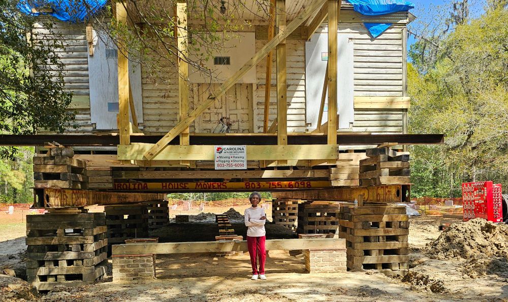 Outside of the rural historic Berkeley County Church, Taveau Church, with a Black woman standing in the front and the church is lifted on stilts. 