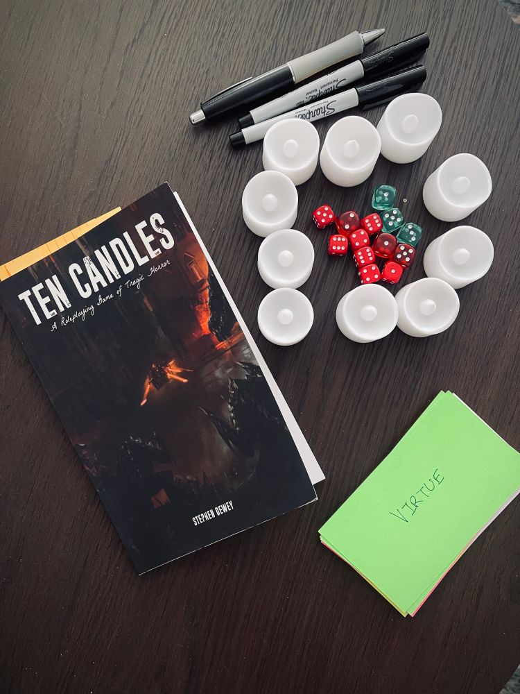 A copy of Ten Candles next to ten LED candles, ten red dice, 3 green dice, a stack of cards (the top one is green and is labeled Virtue) and 3 pens 