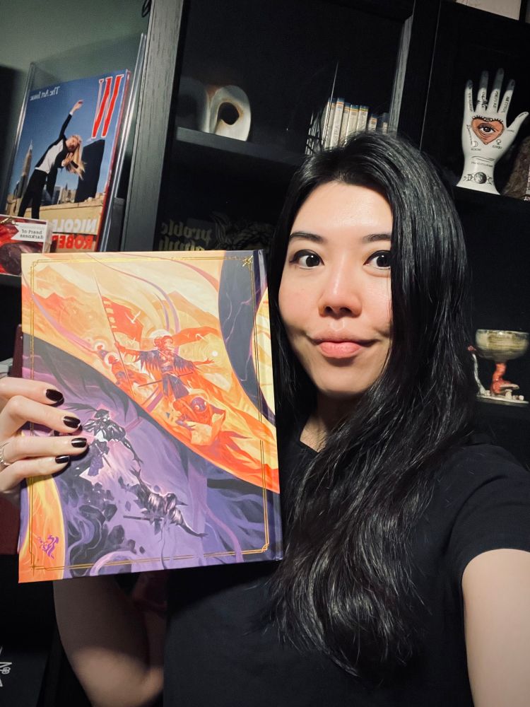 Banana holding up a copy of Daggerheart (it has an orange and purple contrasting cover)