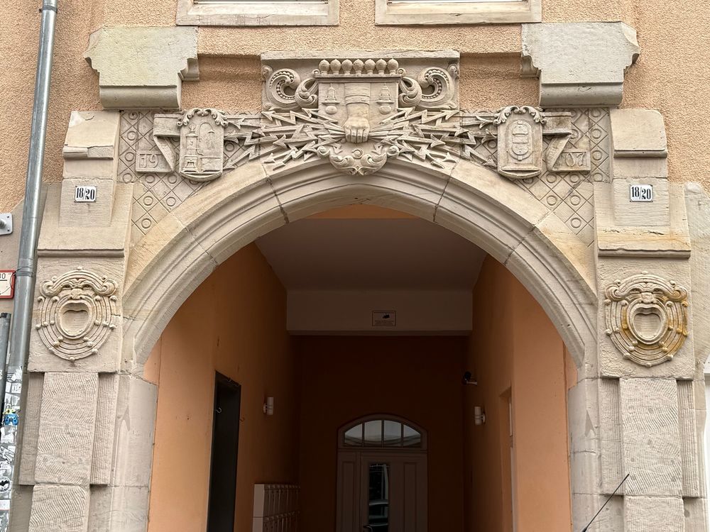 Das Portal in der Detailansicht. Zu sehen ist ein breiter bogenförmiger Eingang ins Haus, darüber mittig eine Hand, die ein Bündel nach rechts und links verlaufender Blitze hält. Rechts und links davon je eine wappenförmige Tafel. Auf der linken ist ein Haus mit einer davor stehenden leuchtenden Straßenlaterne abgebildet.