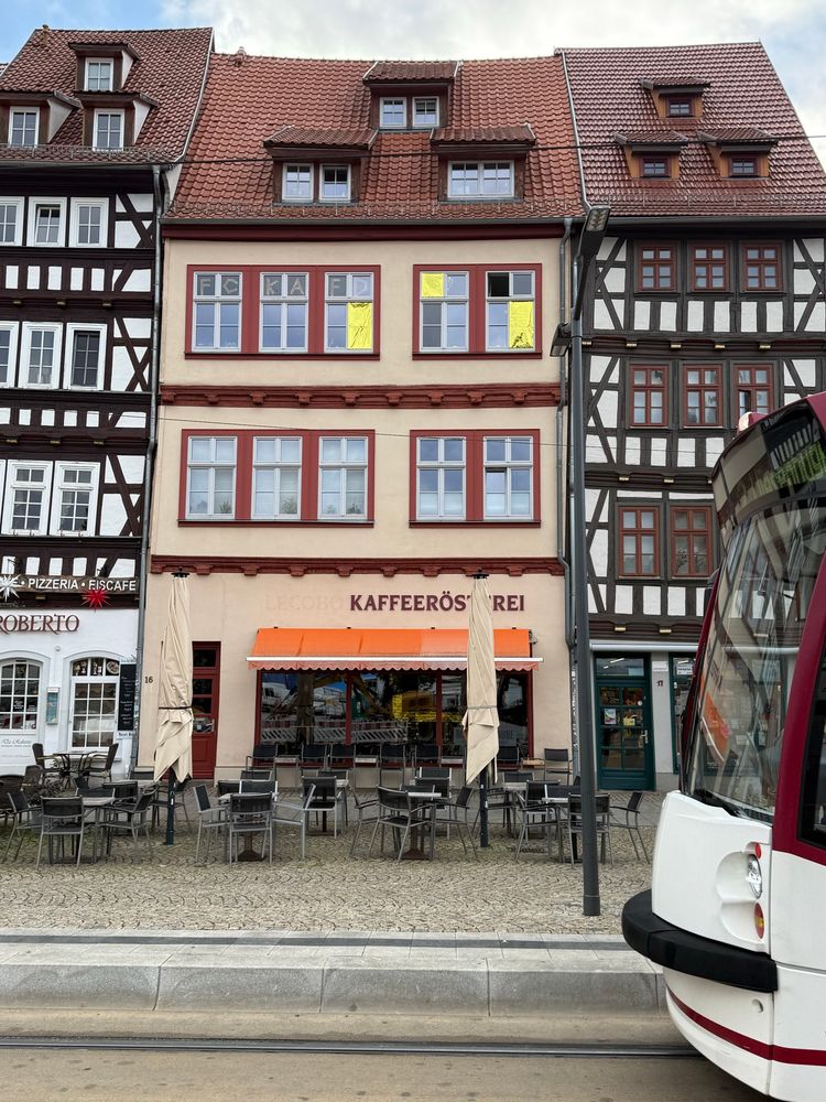 Ein von zwei Fachwerkhäusern eingerahmtes dreistöckiges Gebäude. Im Erdgeschoss eine Gewerbeeinheit, darüber der Schriftzug KAFFEERÖSTEREI. In der oberen Sprossenfensterreihe sind drei Fenster mit goldfarbener Rettungsfolie verhängt. In sechs kleinen Fenstersegmenten wurden die Buchstaben FCKAFD geklebt.