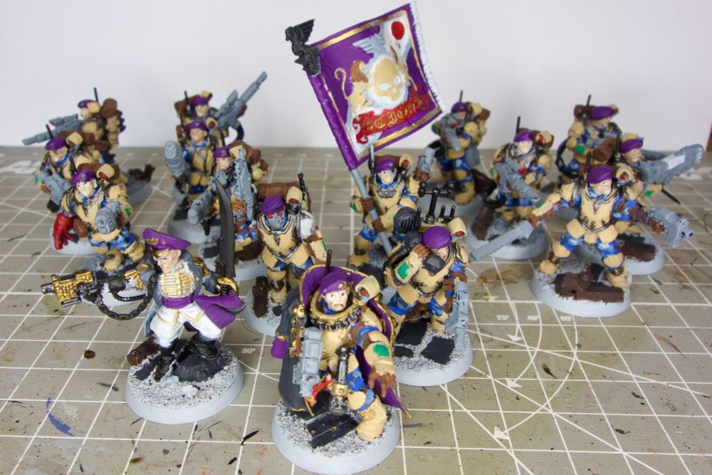 A WIP picture showing 16 Games Workshop miniatures for the game Warhammer 40k. The miniatures are 1 commissar, 1 Militarum Tempestus Command squad, and 2 Militarum Tempestus Scions squads.
The Scions are wearing a blue camouflage uniform, armor in desert tan, and purple berets. Most of their weapons are light grey. The commissar is wearing a grey coat, and a white uniform with golden accents. His hat and his scarf are purple.