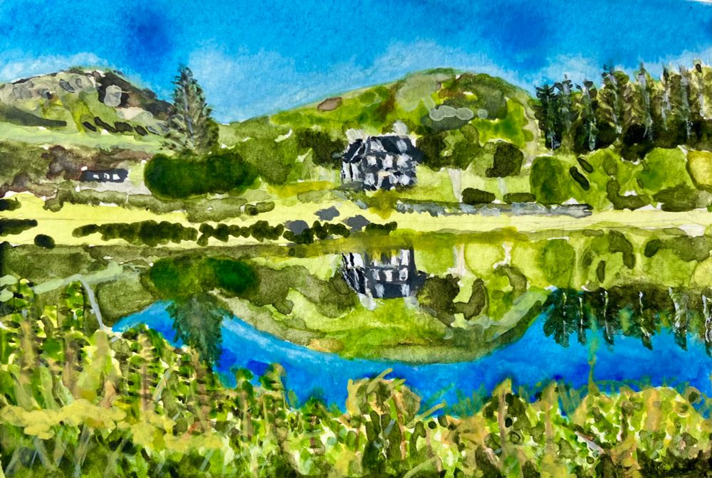 A watercolour painting on a small pocket sized pad. In the foreground are nettles, brambles and long grass that overlooks a former slate mine that is filled with salt water. In the background in the centre is a large, grey stone built house with a backdrop of hills and craggy slopes. There is a large pine tree to the left and more on the far right. The pine trees, hills and house are reflected on the water.