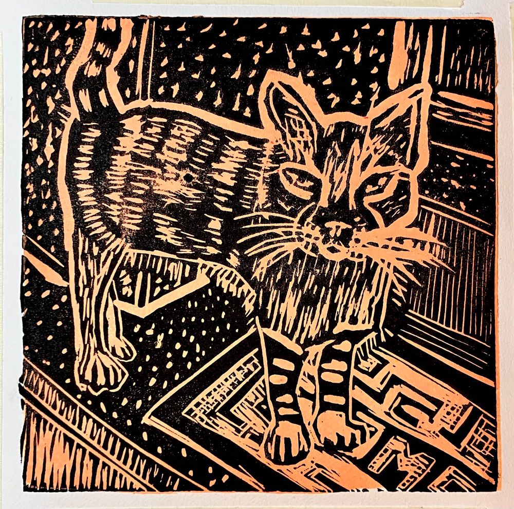 Lino print in peach orange and black of a cat standing on a mat on a raised doorstep in front of a wooden doorway. Behind is a wall. The cat has stripes on its front legs and body. 