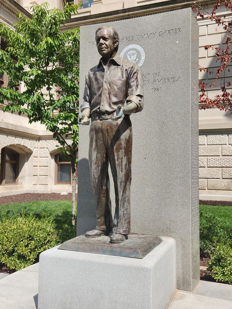 Angled view of the granite and bronze statue depicting Jimmy Carter at the Georgia State Capitol building grounds. The statue depicts President Carter in a button-down shirt with the sleeves rolled up, tucked into plain trousers. His hands are open, palms up. The granite slab behind the statue features the presidential seal and reads "The Honorable Jimmy Carter, 39th President of the United States of America, 1977-1981." 