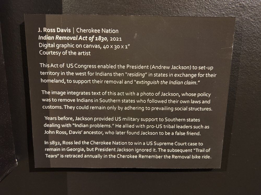Photo of an art exhibit placard that reads:
J. Ross Davis, Cherokee Nation
Indian Removal Act of 1830, 2021
Digital graphic on canvas, 40x30x1"
Courtesy of the artist
This Act of US Congress enabled the President (Andrew Jackson) to set-up territory in the west for Indians then "residing" in states in exchange for their homeland, to support their removal and "extinguish the Indian claim." The image integrates the text of this act with a photo of Jackson, whose policy was to remove Indians in Southern states who followed their own las and customs. They could remain only by adhering to prevailing social structures. Years before, Jackson provided US military support to Southern states dealing with "Indian problems." He allied with pro-US tribal leaders such as John Ross, Davis' ancestor, who later found Jackson to be a false friend. In 1832, Ross led the Cherokee Nation to win a US Supreme Court case to remain in Georgia ,but President Jackson ignored it. The subsequent "Trail of Tears" is retraced annually in the Cherokee Remember the Removal bike ride.
