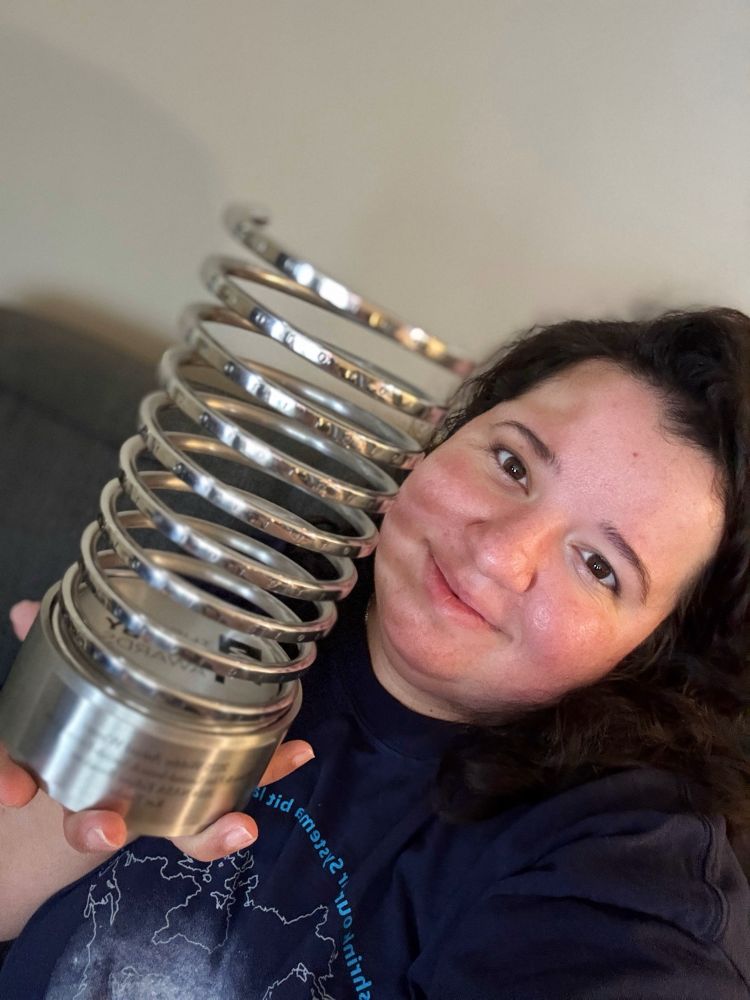 A person smiling while holding a large Webby Award trophy close to their face. The trophy features a shiny silver coil design with engraved binary code and a metal base with an inscription. The person is wearing a dark shirt with a light blue map-like graphic. The background is a neutral-colored wall.