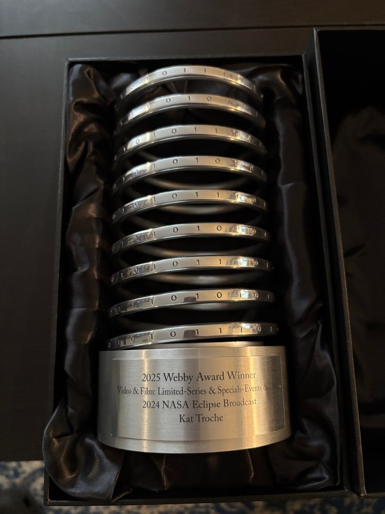 A close-up photo of a 2025 Webby Award trophy nestled in a black satin-lined box. The trophy features a silver coil design engraved with binary code (0s and 1s to spell W-E-B-B-Y) and a metal base with an inscription that reads:
“2025 Webby Award Winner
Video & Film: Limited-Series & Specials–Events & Live Streams
2024 NASA Eclipse Broadcast
Kat Troche.”