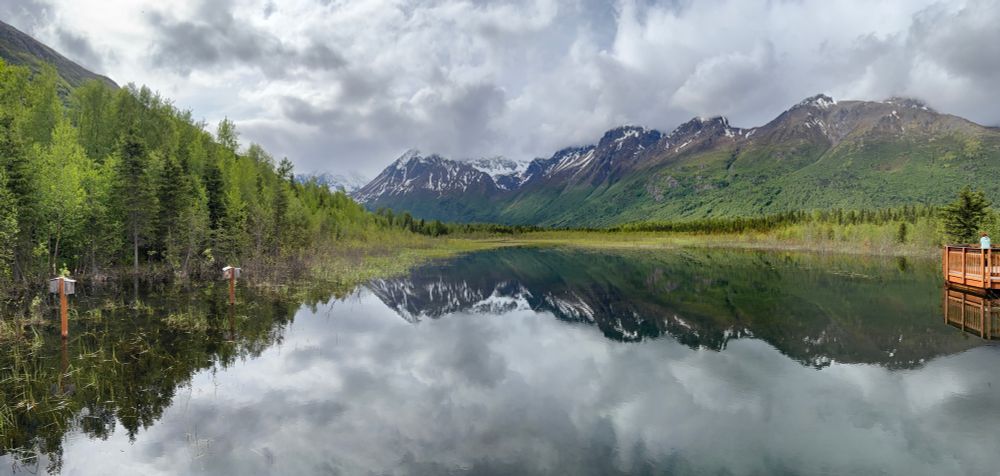 A dark mountain range with snow covering the tops of the mountains in the background, with a dense green forest in the mid ground, and a lake in the foreground. The mountains are reflected in the lake due to its clarity. 