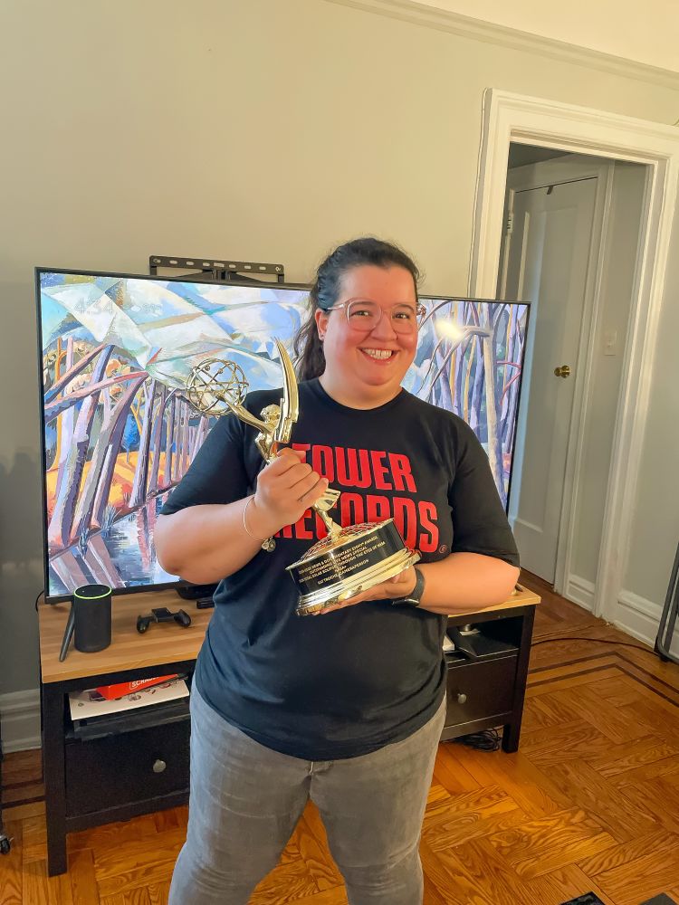 A smiling woman stands in a living room holding an Emmy® Award. She is wearing glasses and a black t-shirt with bold red text that reads “TOWER RECORDS.”