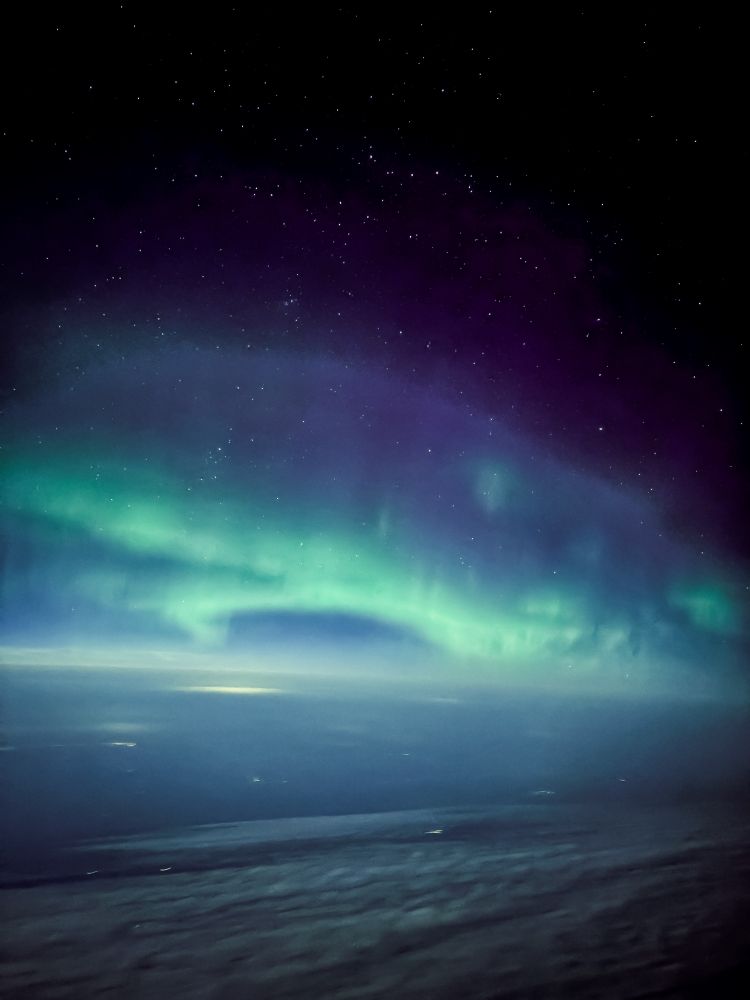Swirling green bands of Aurora in the night sky above a thick band of clouds, as seen from an airplane 30,000 feet up. Near the top of the aurora, purple colors appear along with a dark night sky scattered with stars.