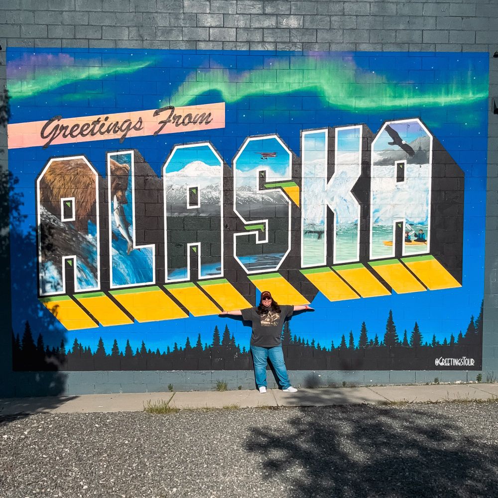 A woman in blue jeans, a gray shirt and a red ball cap standing in front of a mural pained on the side of a building that says 'Greetings from Alaska' with the northern lights painted into the picture, and various scenes of a bear catching salmon and an eagle flying over mountains and a glacier painted into the letters.