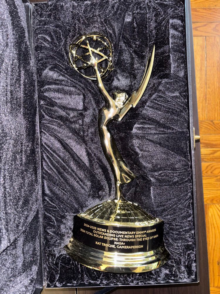 A close-up of an Emmy® Award resting in a black velvet-lined display case. The gold statue features a winged figure holding an atom. The base inscription reads:
2024–2025 News & Documentary Emmy® Awards
Outstanding Live News Special
2024 Total Solar Eclipse: Through the Eyes of NASA
NASA+
Kat Troche, Cameraperson