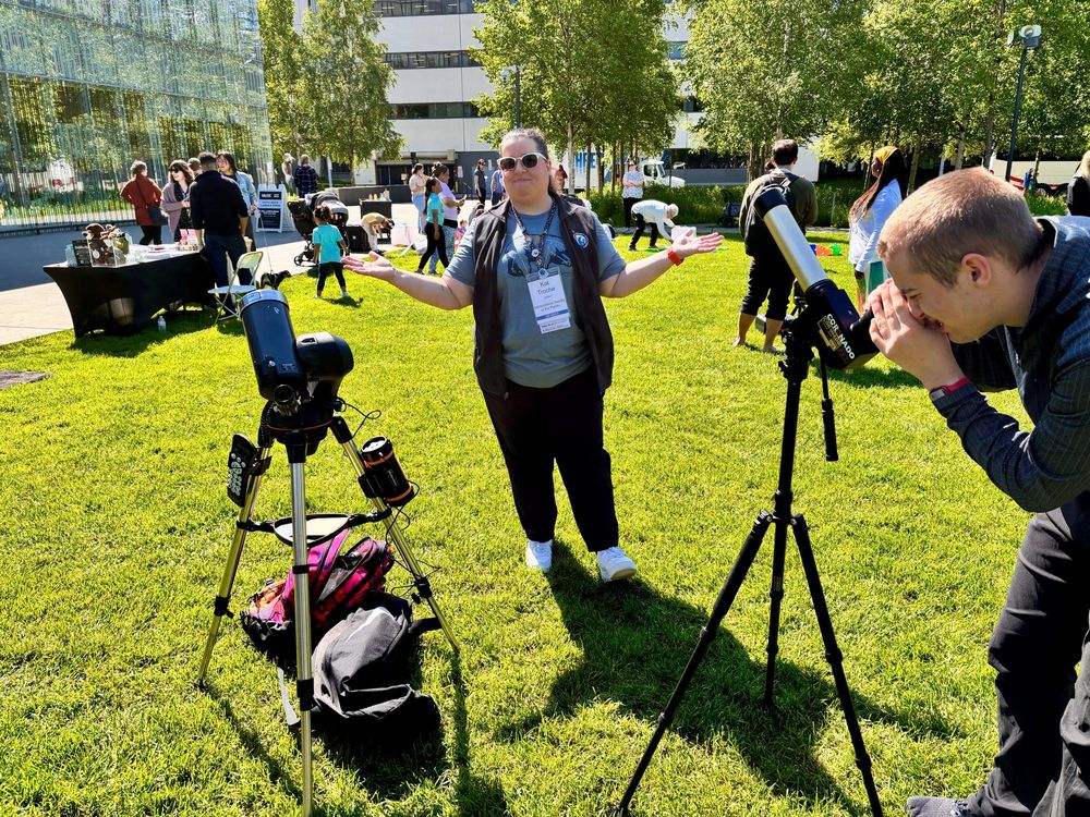 A woman standing on a lawn with green grass, between two telescopes that are looking at the sun, and a person looking through one of telescopes. There are several people in the background walking around.