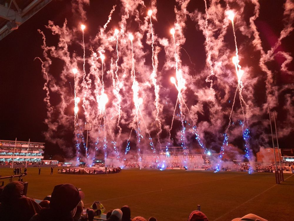 Red fireworks at night are launched from the pitch at Belle Vue stadium in Wakefield, with stands in the background and spectators in the foreground. 