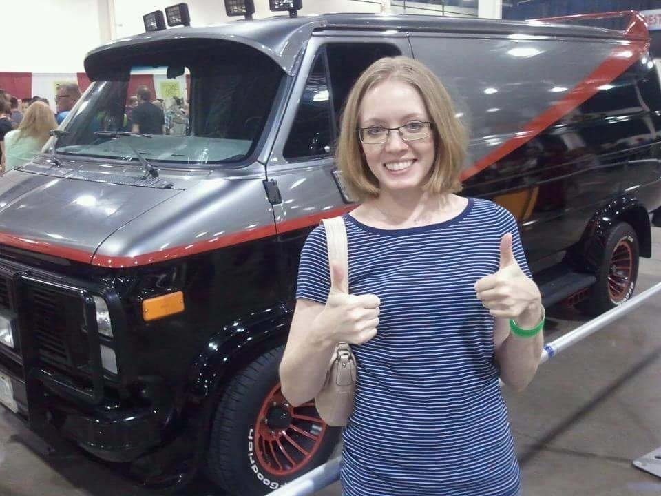 A smiling woman in a striped shirt and glasses gives a double thumbs up while standing in front of the A Team van