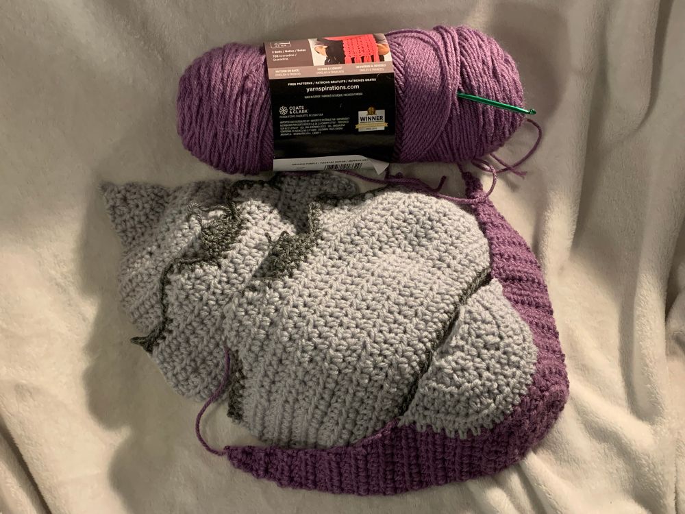 A photo of an unfinished, life-sized-ish crocheted wimpod Pokémon. It has its shell, but is missing its tail, legs, belly, and face so far. It is sitting next to a purple skein of yarn.