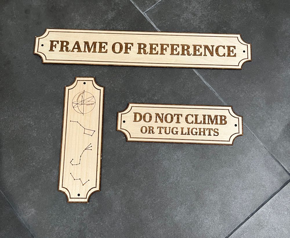Laser cut and etched wood plaques made in a sort of vintage Victorian aesthetic. One reads “FRAME OF REFERENCE”, another “Do not climb or tug lights”, and the last has pictures of the constellations along with a diagram of celestial coordinates. 