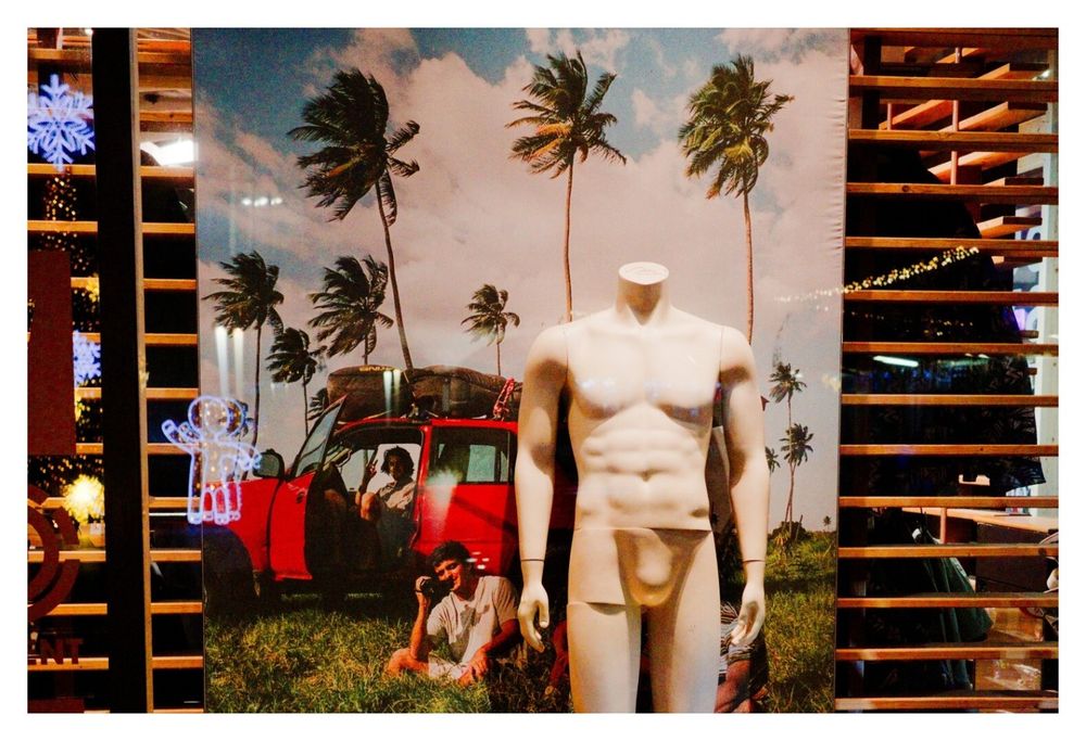 vitrine de magasin de vêtements, mannequin masculin sans tête, en fond une photo de jeunes gens sortant d'une voiture rouge sous des palmiers, l'un nous film l'autre nous fait signe de la main