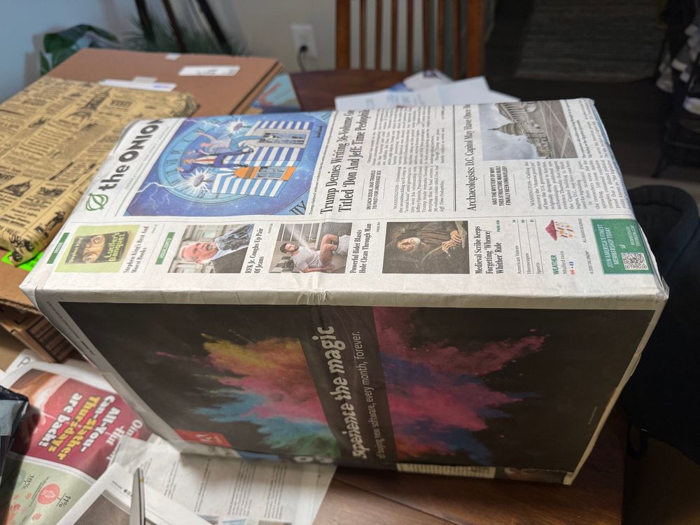 Box wrapped with The Onion newsprint. Some of the headlines read “Trump denies writing 36-Volume comic titled ‘Don and Jeff: Time Pedophiles”, RFK Jr coughs up a pair of jeans, powerful bidet blasts hole clean through man, medieval scribe keeps forgetting ‘whence/whither’ rule, Archaeologists: DC may have once been used for legislating. A full page “not a paid advertisement” for Adobe on one side of the box reads “experience the magic of buying new software, every month, forever.”