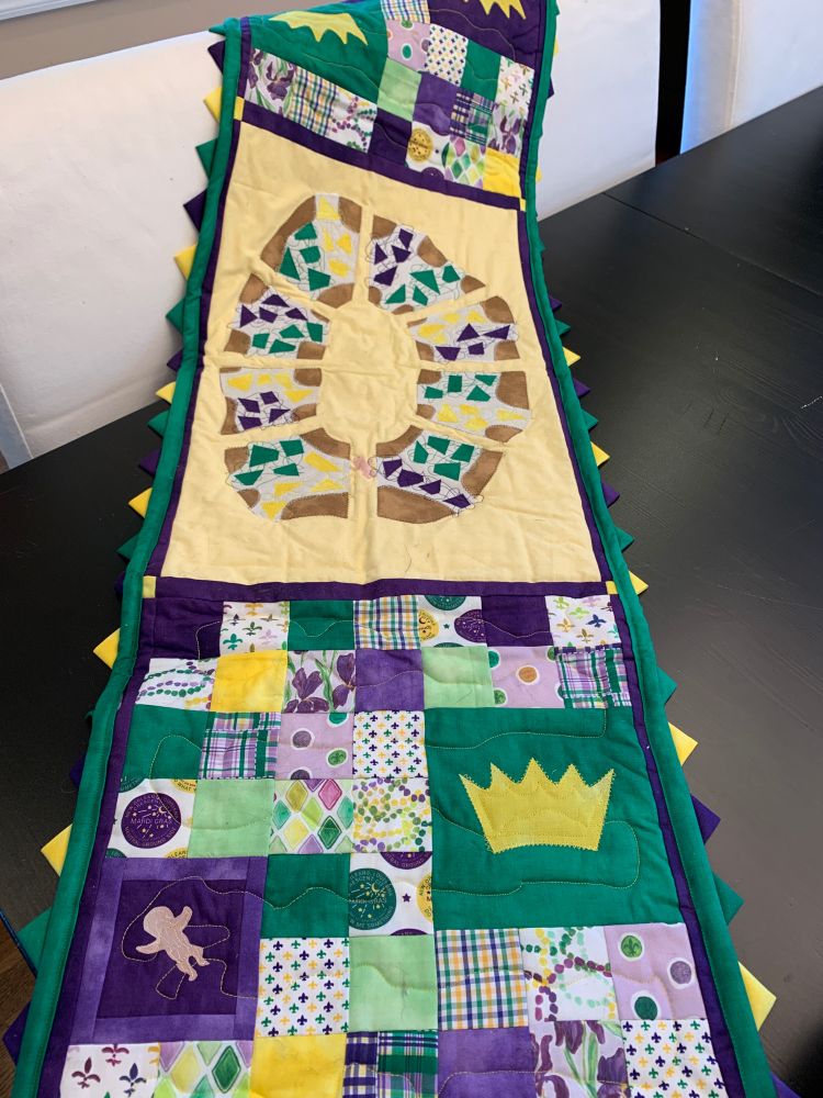 Photos of a table runner in colors of purple, green, and yellow. In the center is a yellow field with a design of a king cake on top. On the ends are squares of various patterned fabric.