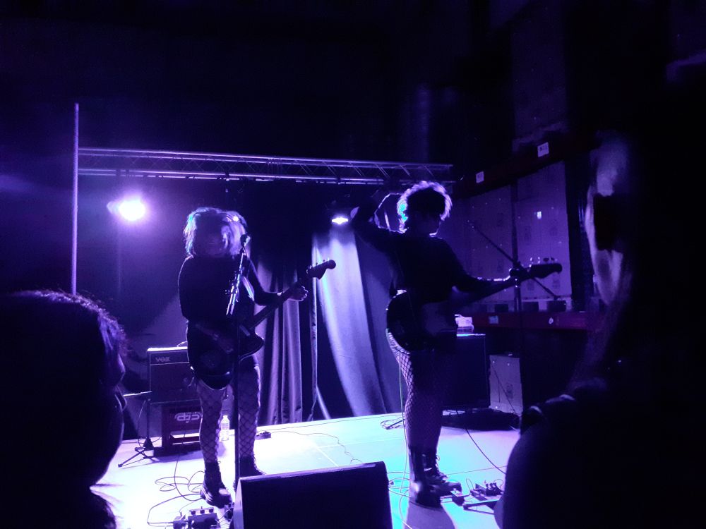 Diavol Strain playing on a scene with purple lights ; the guitarist, Ignacia Strain, is on the left ; the bassist and vocalist  Lau M, is on the right ; they're close to each other and beautiful 