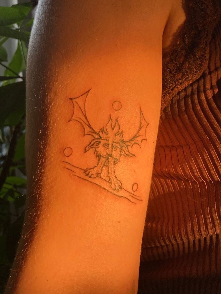 Tatouage d’un grotesque médiéval, une tête d’un genre de creature poilue avec des ailes de chauve souris et des pattes de lion dans sa bouche. Il est posé sur une branche avec trois orbes qui l’entourent. Goofy guy pris en photo pendant la golden hour avec une belle lumière chaude 