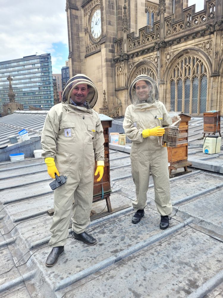 Two people in bee suits on a roof with wooden hives behind