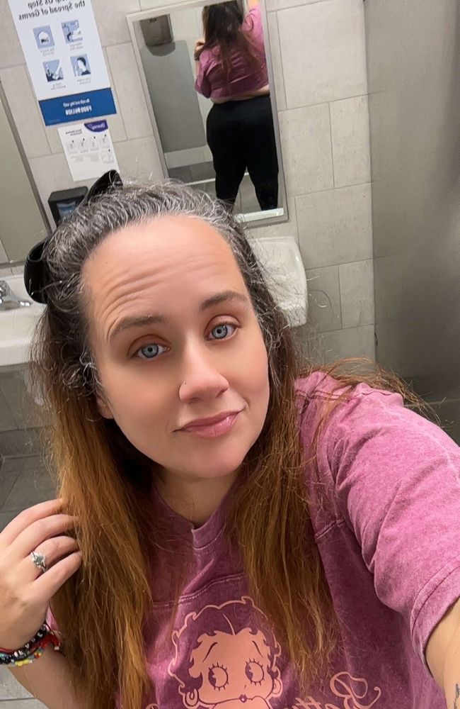 Pictured is a thick, brunette with blue eyes that’s looking at the camera, her right hand in her hair. She has her hair pulled back but let down and it goes past her shoulders. Shes wearing a mauve colored Betty boop graphic tee that’s been cut into a crop top. Behind her there’s a mirror showing the back, her thick ass in tight black leggings. She has jewelry on. 