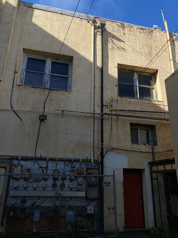 Photo of the side of a yellowing building. There are cables and pipes and electrical meters on the side of the building. A triangle of sunlight and blue sky can be seen in the top right corner of the image.