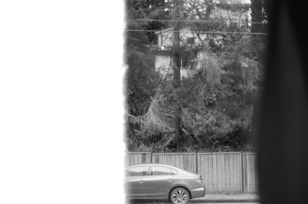Black and white film photo of a car shot through a window. Behind the car lies a fence, trees, and a house on a hill. The car's front half is bisected by a jarring white bar that takes up about a third of the frame, beginning on the right. This white shape is the overexposed white curtain being illuminated by the light. 