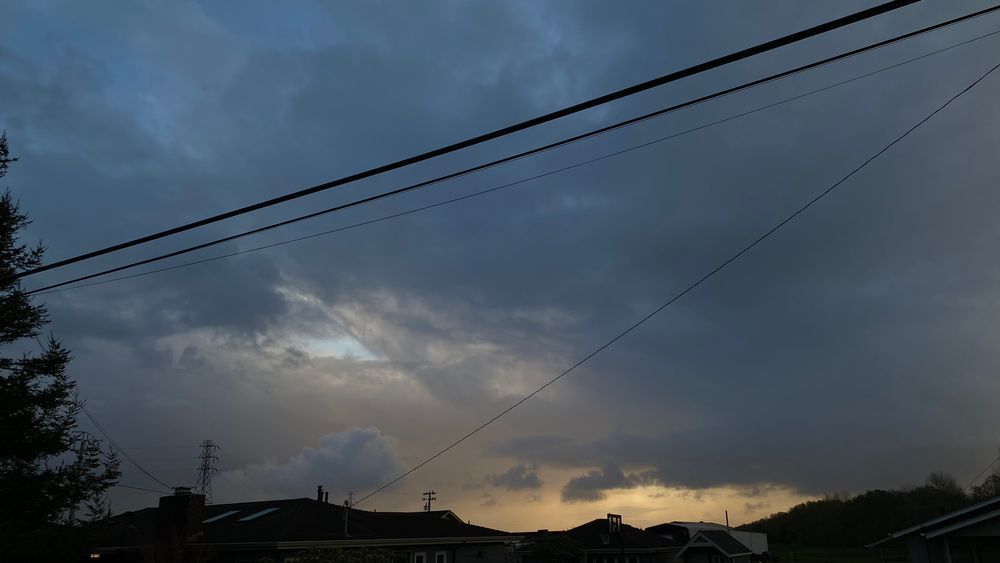 Photo of a stormy sky with grays, blues, and oranges lighting up the cloudy background. Telephone wires and the tops of houses can be seen. 