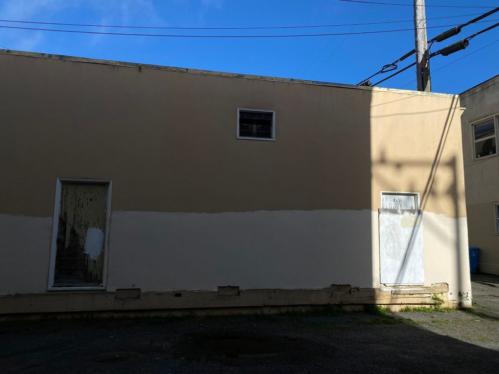 Side of a yellow-beige building that is losing its luster with boarded up doors. Light shines on the right side of the building, and the blue sky can be seen overhead.