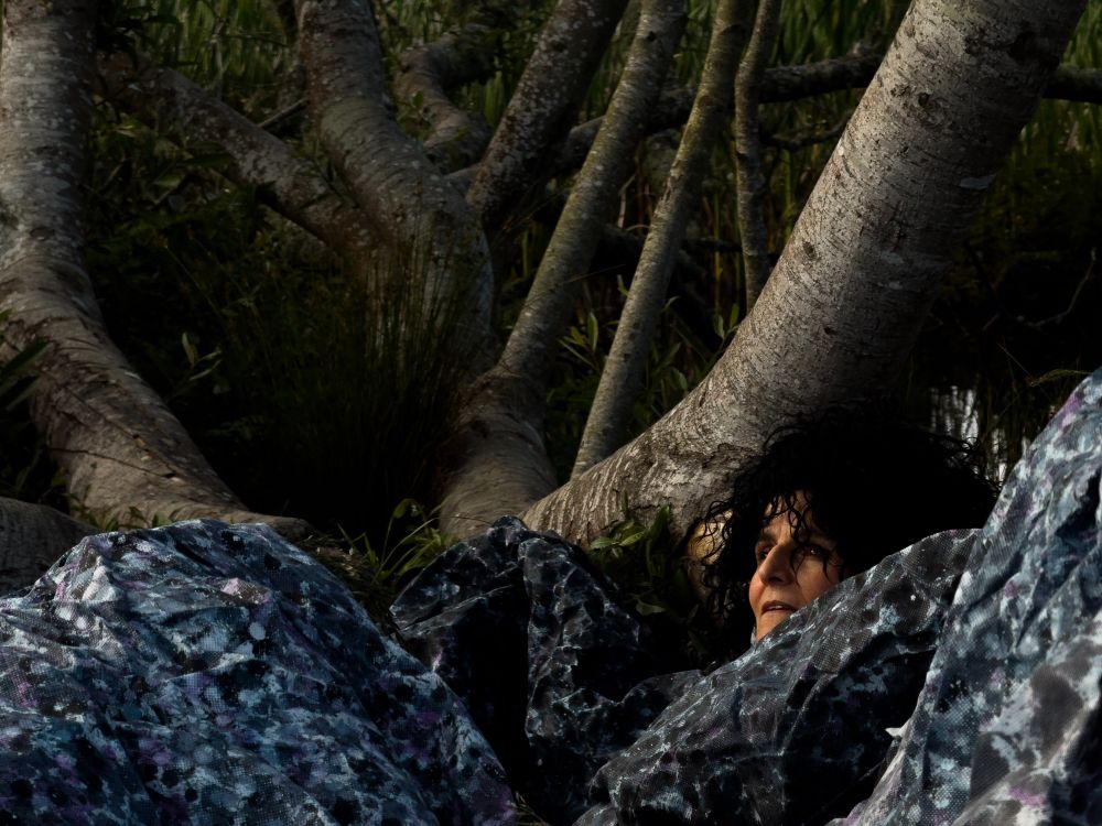 Photograph of a distressed-looking woman. She is laying down under a tree, her gaze affixed upwards. She is covered in some kind of tarp or blanket covered in a dark pattern. The reflection of water in a pond reflects some light behind her.