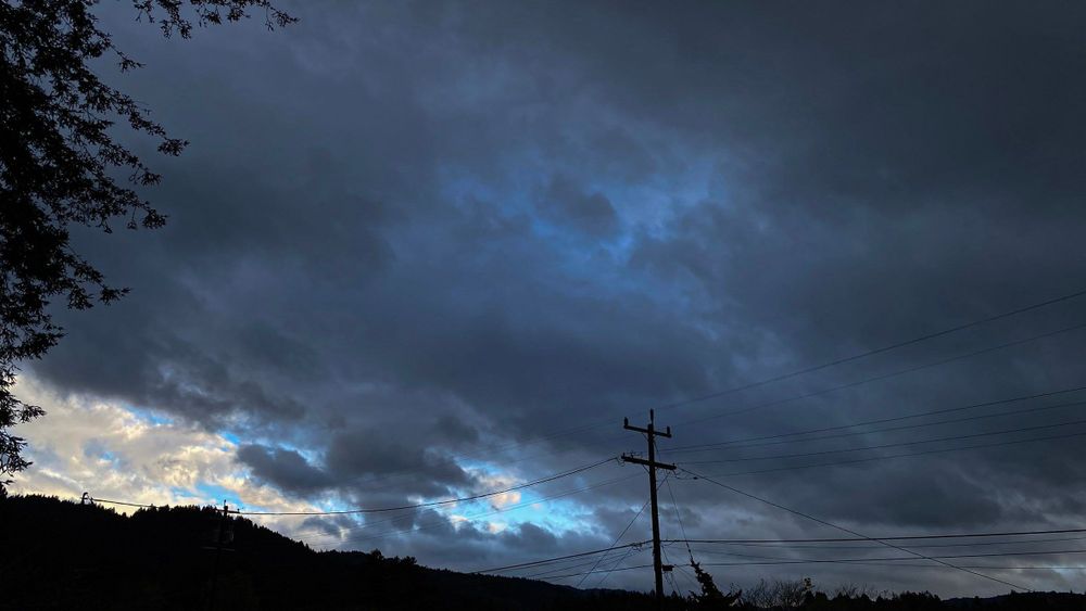 Color photograph of a stormy and cloudy sky. Blue streaks and lit-up white puffy clouds can be seen in the bottom left corner of the image. A telephone pole sits squarely in the middle of the shot and forested mountains can be seen in the background. 